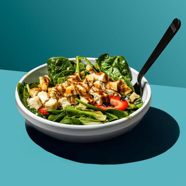 A vibrant spinach salad with grilled chicken, strawberries, and balsamic glaze in a white bowl.