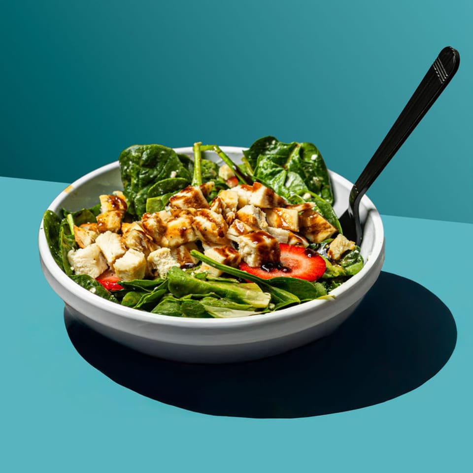 A vibrant spinach salad with grilled chicken, strawberries, and balsamic glaze in a white bowl.
