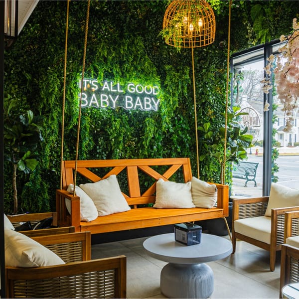 Good Life Clubhaus interior with a wooden swing, green wall, and "IT'S ALL GOOD BABY BABY" neon sign.
