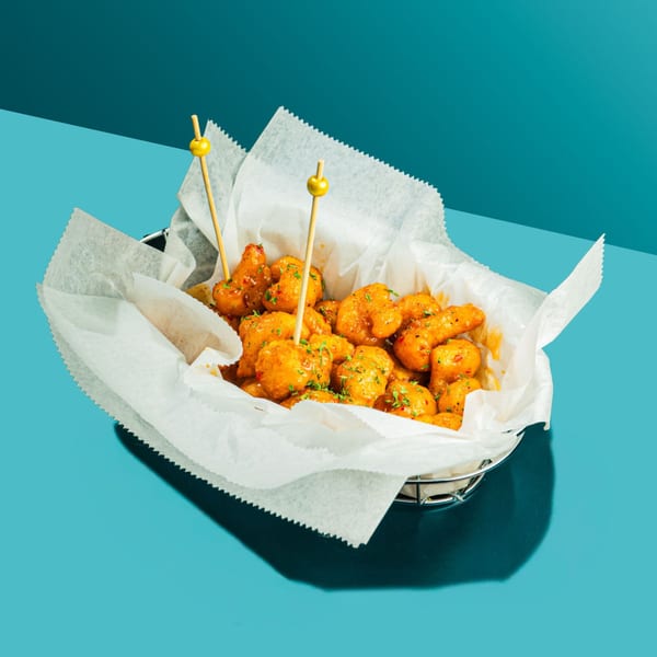 A wire basket filled with crispy orange cauliflower bites, garnished with green herbs and yellow picks.