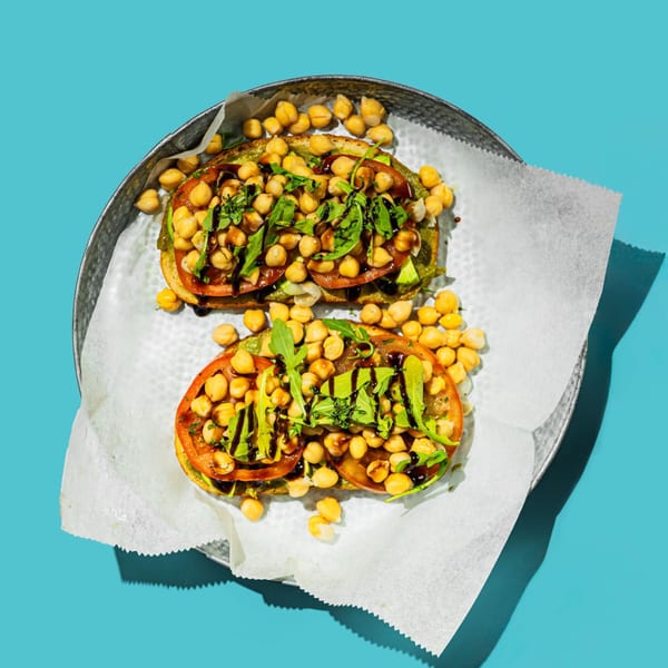 Two avocado toasts topped with chickpeas, tomatoes, arugula, and balsamic glaze on a metal tray.
