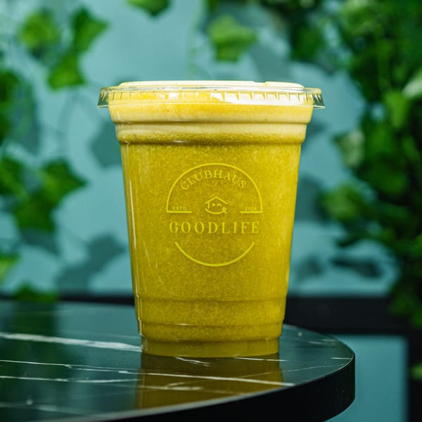 A Good Life Clubhaus green juice in a clear plastic cup with a lid on a marble table.