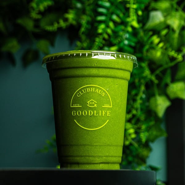 A vibrant green smoothie in a clear cup with the Good Life Clubhaus logo, against a leafy green background.