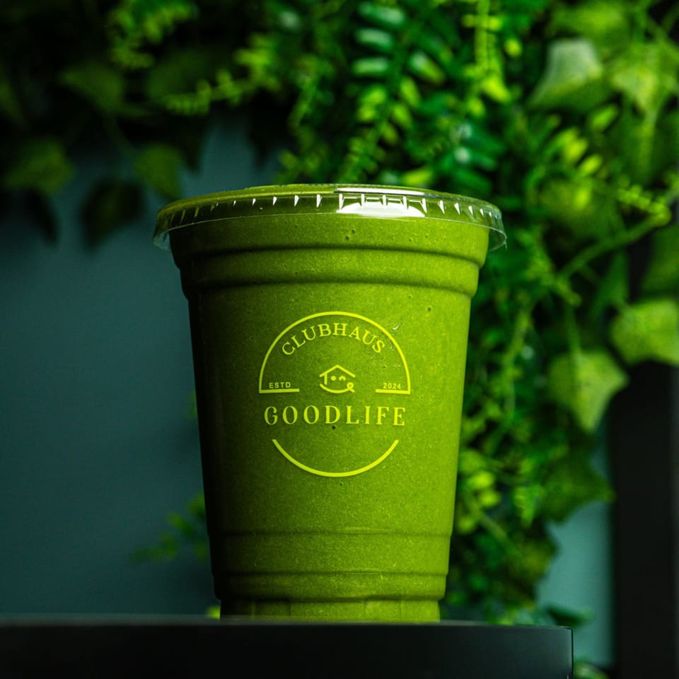 A vibrant green smoothie in a clear cup with the Good Life Clubhaus logo, against a leafy green background.